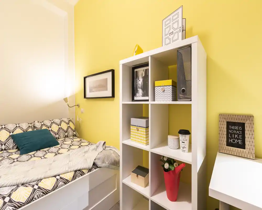 Small, bright bedroom with patterned bedding, a teal accent cushion and a white cube shelf used as storage and decor — warm yellow walls create a cozy atmosphere.