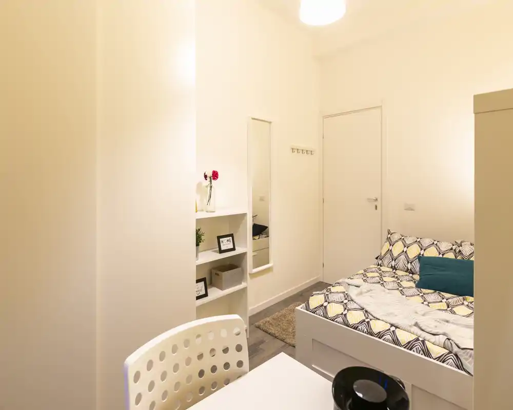 Cozy, well-lit bedroom featuring a patterned bed with cushions, shelving for storage and a small desk area — ideal for a comfortable stay.