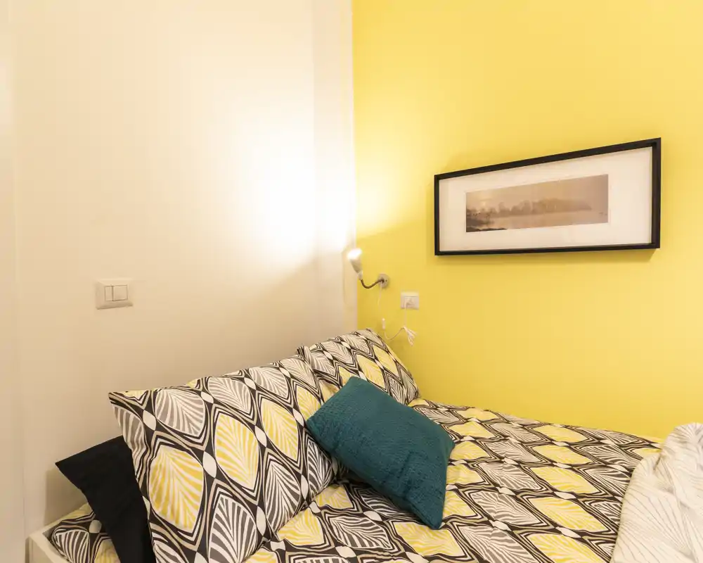 Bright, cozy bedroom with patterned bedding and a teal accent cushion against a sunny yellow wall; warm bedside light and framed art create a welcoming atmosphere.