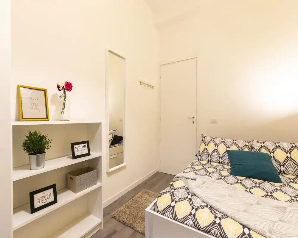 Bright, well-staged bedroom with patterned bedding, a teal accent pillow, wall mirror and decorative shelving — ideal for a rental listing highlighting a cozy small bedroom.