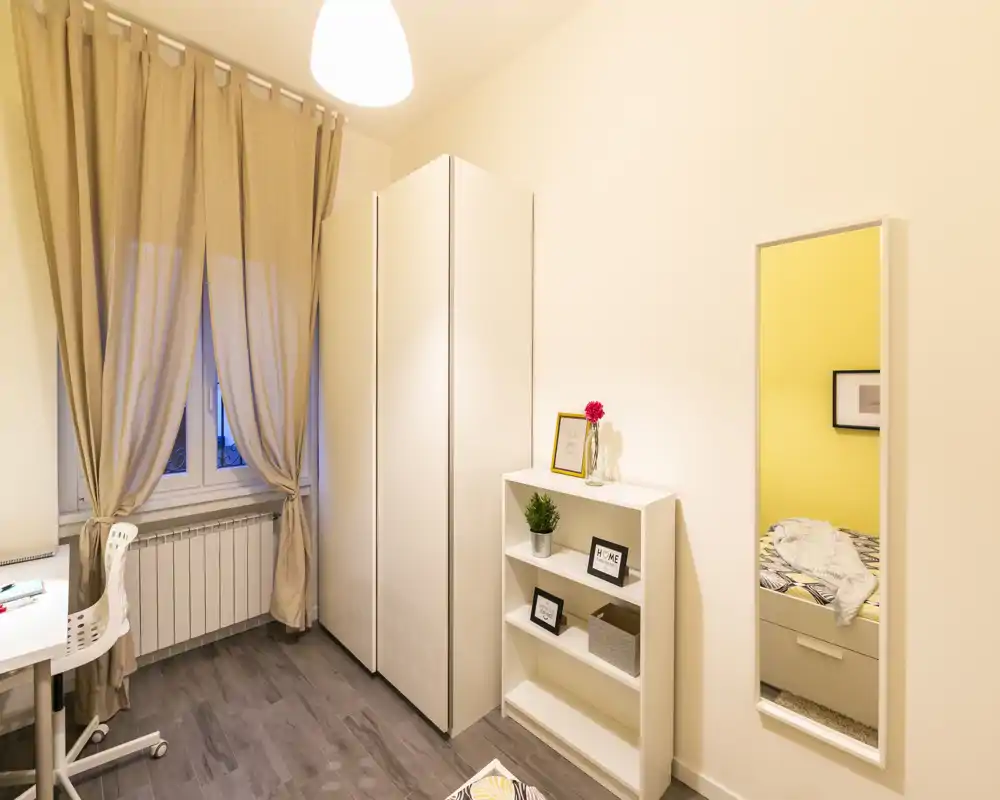 Cozy small bedroom corner with a wardrobe, shelving unit and a wall mirror that shows the bed in reflection. Warm, tidy styling and soft lighting create an inviting atmosphere ideal for a rental listing.