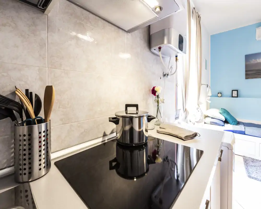 Compact modern kitchenette with an induction hob, cooking utensils and a bright window opening onto a small sleeping area; clean, practical and well lit.