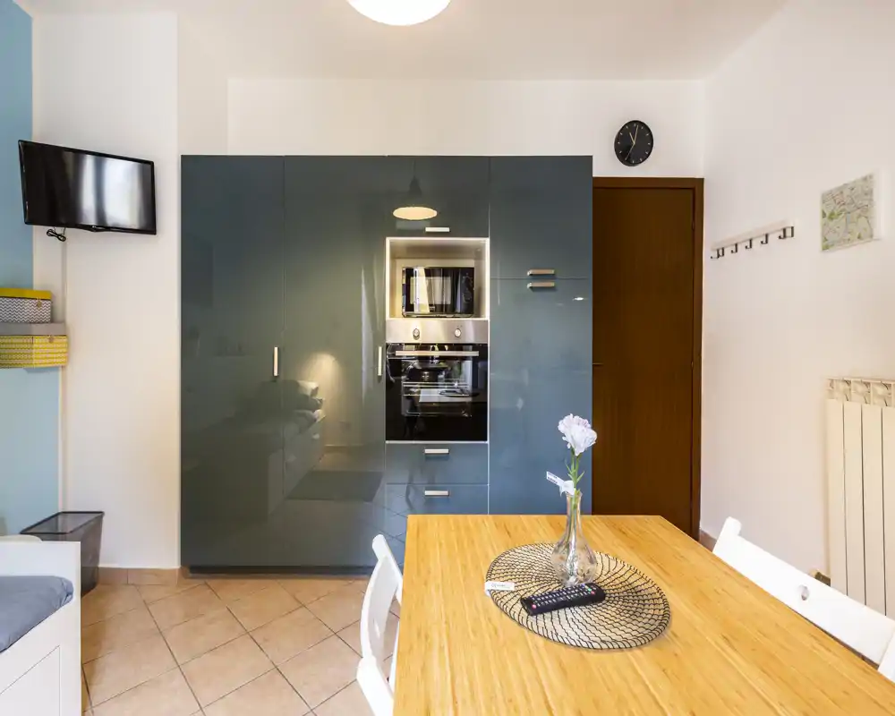 Compact modern kitchen area with glossy built-in cabinetry, integrated oven and microwave, and a wooden dining table with seating — bright, tidy and suitable for short-term rentals.