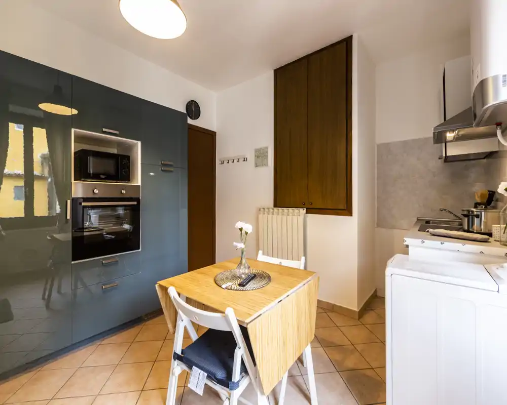 Compact modern kitchen with built-in oven and microwave, a small foldable wooden dining table and practical countertop appliances. Bright, neatly arranged space suitable for short-term rentals.