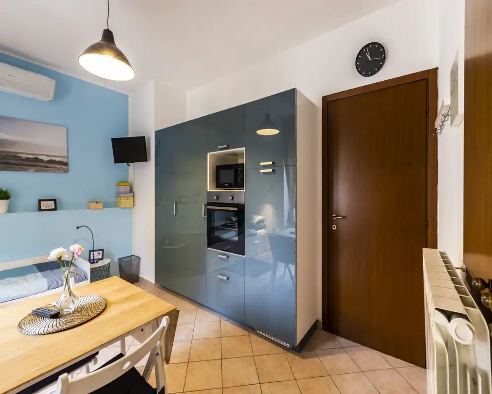 Compact modern kitchen area with integrated glossy cabinets, built-in oven and microwave, small dining table and pleasant decorative touches — ideal for a studio apartment.
