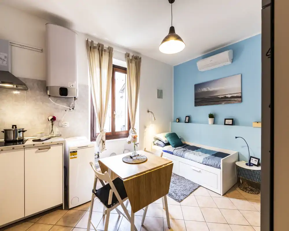 Compact studio bedroom with a single daybed, a small dining table and an integrated kitchenette. Bright natural light, soft blue accent wall and modern, cozy decor ideal for a solo stay.