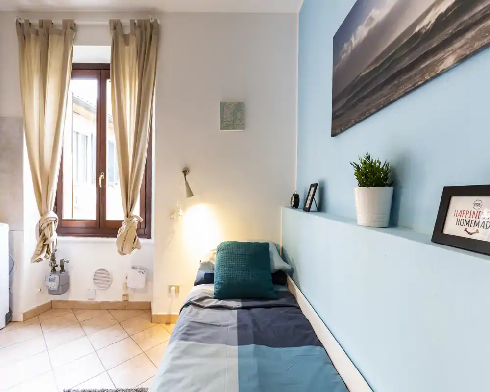 Small, bright single bedroom with a neatly made bed, teal accents and framed decor on a soft blue accent wall. Natural light from a large window, compact layout ideal for a cozy rental.