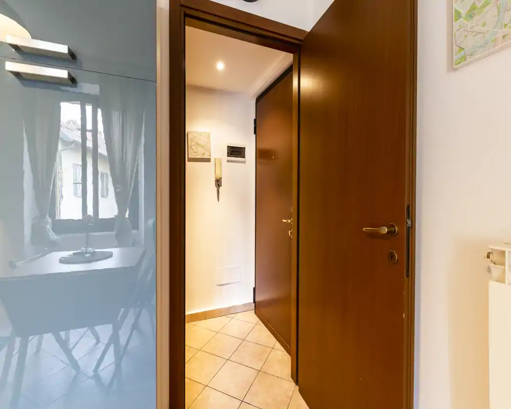 Compact and well-lit entrance hallway with tiled floor and wooden door, showing a clear view into the flat and a nearby dining area reflected on a glossy surface.