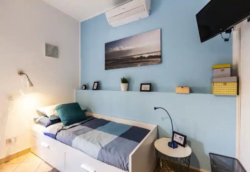 Bright single bedroom with a neat single bed, blue accent wall, wall art and compact bedside table — cozy, well-lit and tastefully decorated for a short-stay rental.