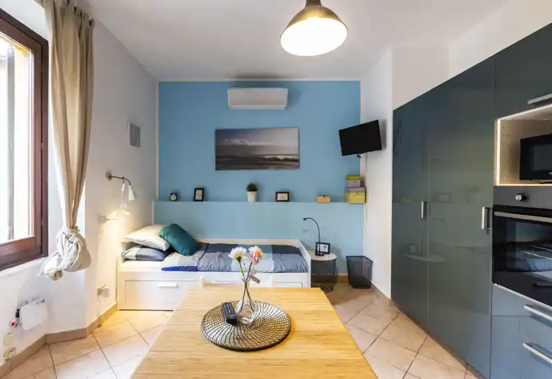 Bright, compact bedroom with a single daybed, soft pillows and coordinated blue accent wall; modern built-in storage and a small dining/work table create a functional studio layout.