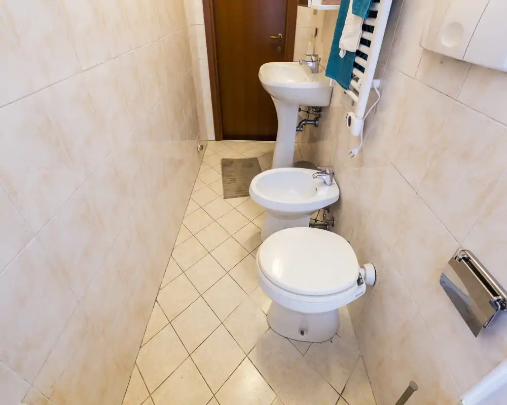 Clean, narrow bathroom with toilet, bidet and pedestal sink arranged along the right wall; neutral beige tiles and good lighting.