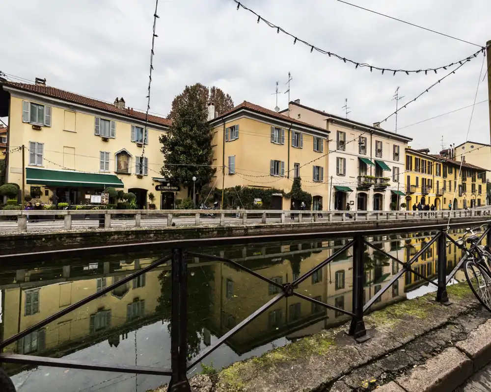 Canal-side view with classic yellow buildings reflected in calm water, a pedestrian railing in the foreground and string lights overhead; atmospheric overcast sky.