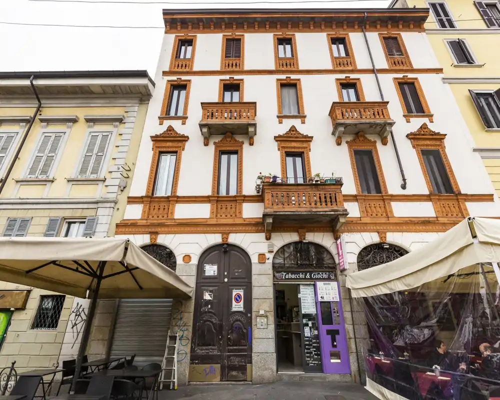 Street-facing building facade with ground-floor shops and outdoor seating under umbrellas; clear view of architectural details and balconies.