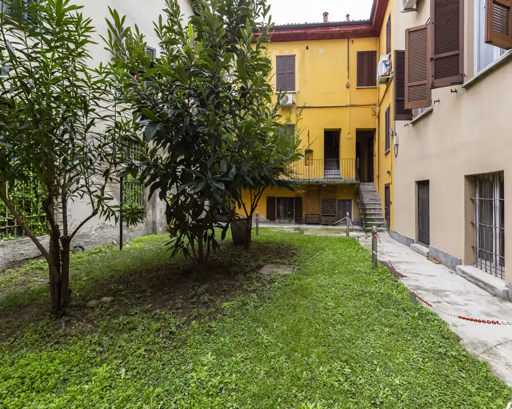 Sunny inner courtyard with a grassy area and small trees, bordered by residential buildings with yellow façades and shuttered windows.