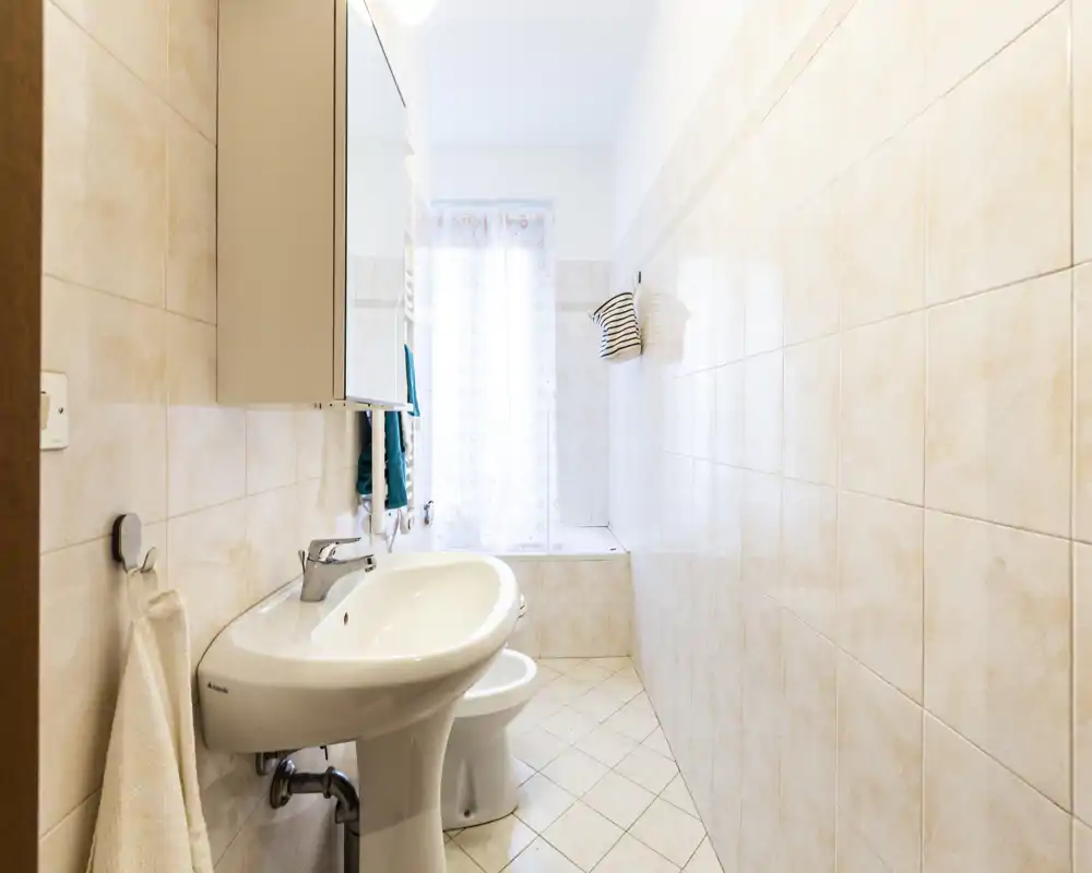 Bright, tidy bathroom with a pedestal sink, bidet, bathtub by the window and light beige tiled walls and floor.