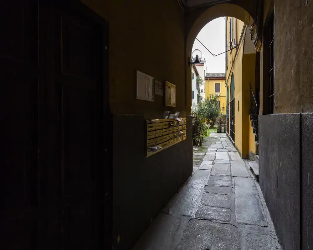 Covered passageway/entrance with stone paving, mailboxes on the left and an arched opening leading to a sunny courtyard beyond.
