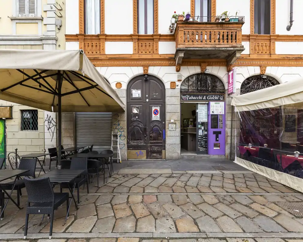 Outdoor café/terrace area in front of a building with tables, chairs and large umbrellas — suitable for al fresco dining or a bar entrance.