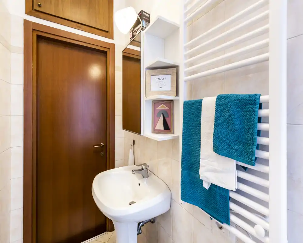 Bright, well-kept bathroom with a pedestal sink, wall-mounted towel radiator and teal towels; neutral tiles and wooden door add warmth.