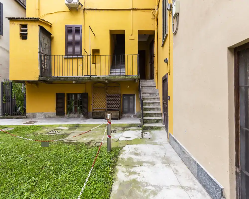Sunny yellow courtyard with stone walkways, a small grass patch and external stair leading to upper doors — a typical shared outdoor space in an older building.