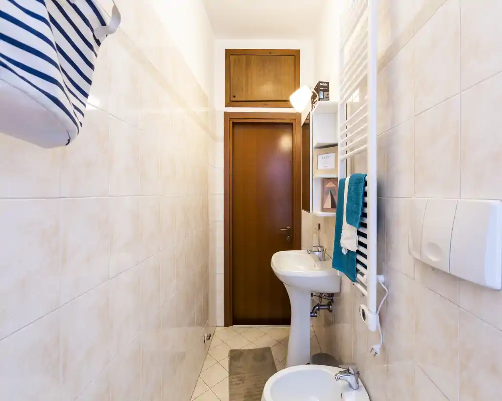 Compact, well-lit bathroom with a pedestal sink, bidet, towel radiator and tiled walls — neat and ready for guests.