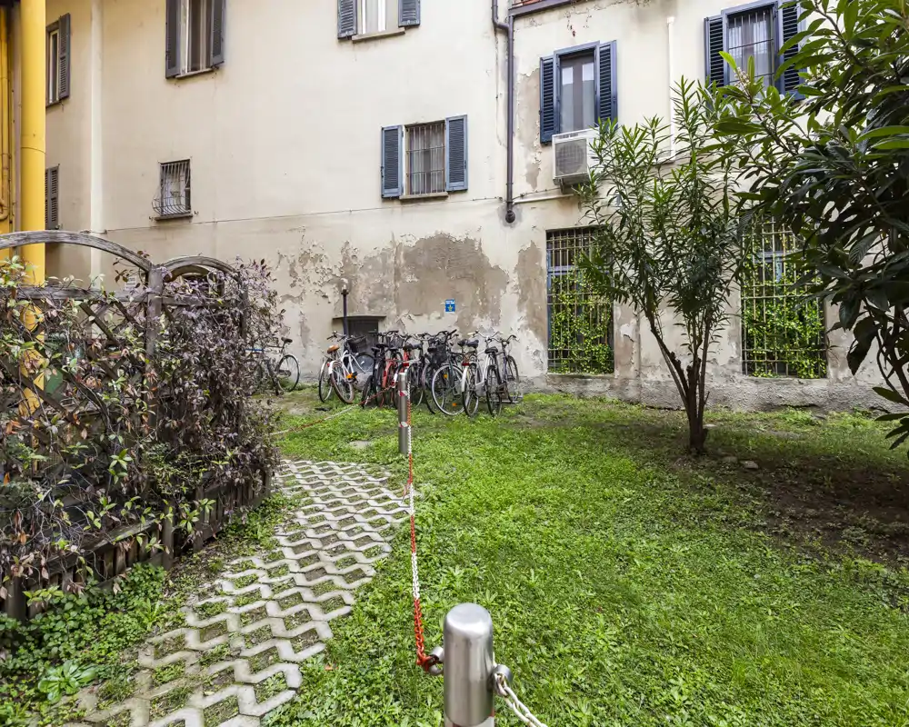 Small private courtyard with grass, a paved path and a parked row of bicycles against a building wall. Green shrubs and windows with shutters give it a quiet, urban-garden feel.