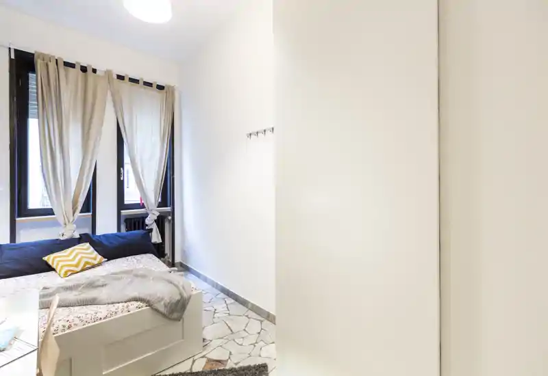 Camera da letto luminosa e dallo stile semplice con un letto matrimoniale vicino a grandi finestre che lasciano entrare molta luce naturale. Pareti neutre, biancheria con fantasia e un plaid rendono l'ambiente accogliente.