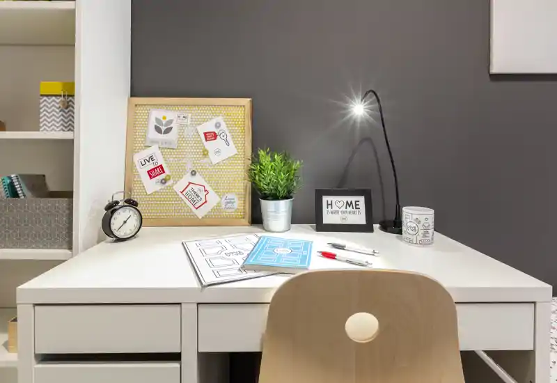 Angolo studio moderno e luminoso con scrivania bianca, lampada da tavolo, piantina e bacheca — ideale per smart working o studio.