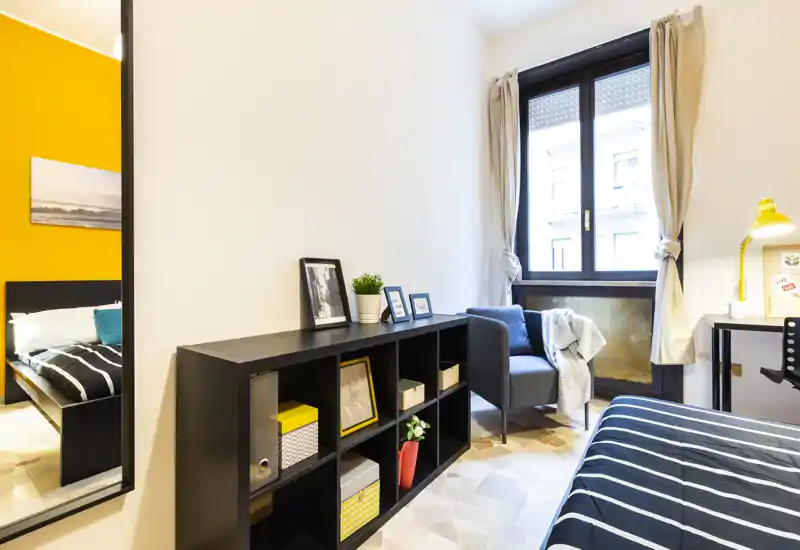 Camera da letto luminosa e ben arredata con letto a righe, mobile contenitore, poltrona accogliente vicino alla finestra e una scrivania per lavorare.