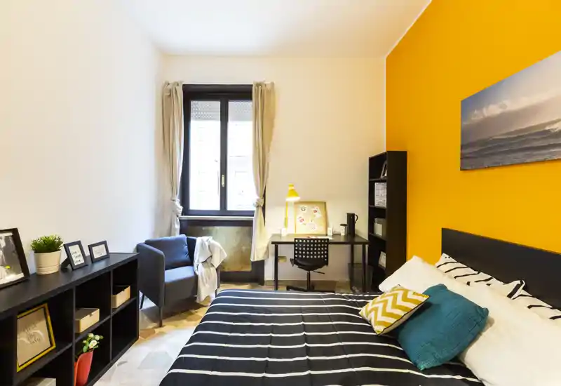 Camera da letto luminosa e ben arredata con un comodo letto matrimoniale, cuscini decorativi e una parete arancione vivace. Comprende una scrivania vicino alla finestra e una poltrona accogliente, perfetta per un annuncio di affitto.