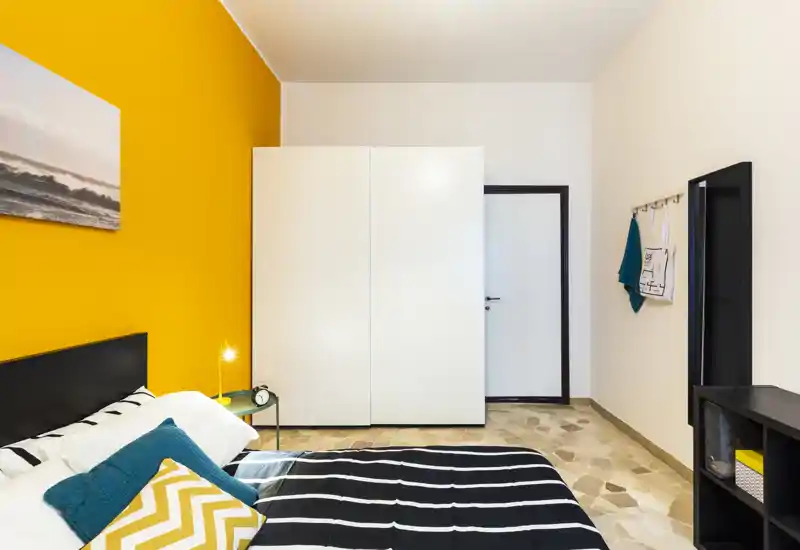 Camera da letto moderna e luminosa con parete d'accento gialla, letto ordinato con biancheria a righe e cuscini colorati, armadio bianco e arredamento minimale.