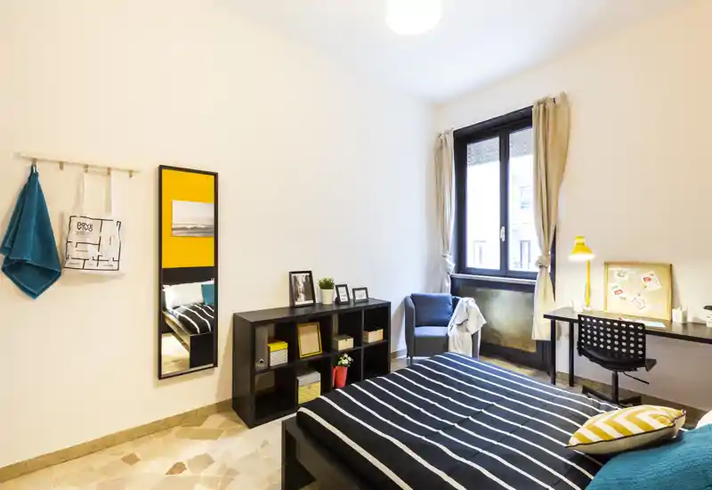 Camera da letto luminosa e moderna con letto a righe, libreria a cubi, scrivania e finestra che illumina la stanza. Arredo semplice e curato, con cuscini e angolo studio ideali per un annuncio di affitto.