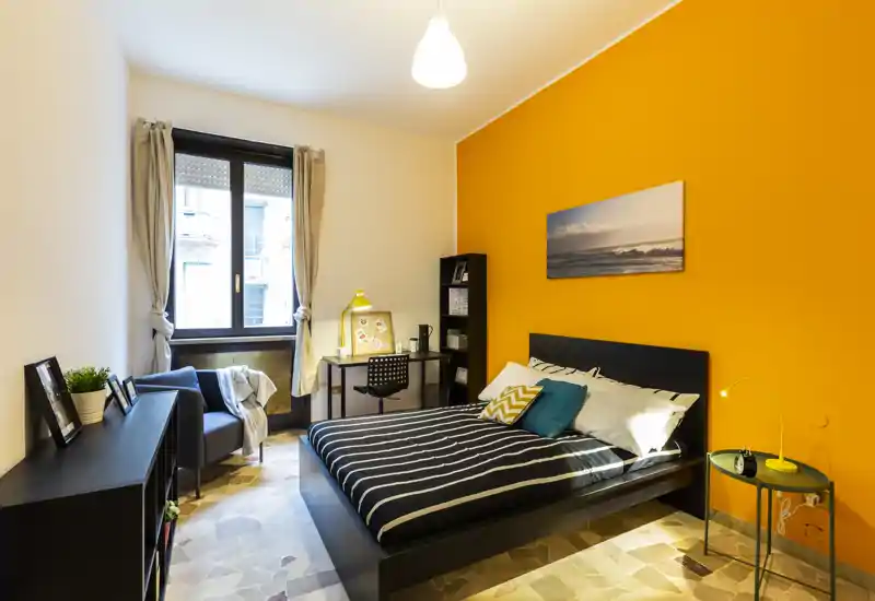 Camera da letto luminosa e di design con letto matrimoniale davanti a una parete arancione, comodino con lampada, scrivania e poltrona vicino alla finestra — moderna, accogliente e ben illuminata.