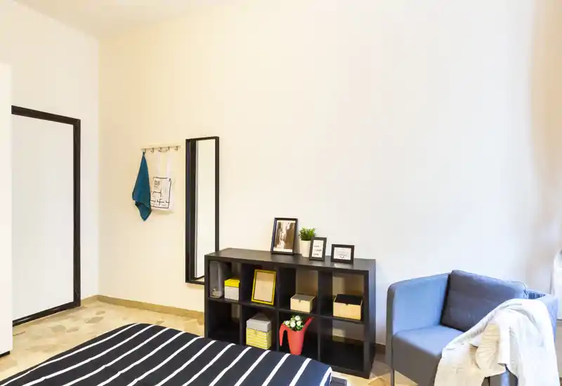 Camera da letto luminosa e ben allestita con letto a righe in primo piano, scaffalatura con oggetti decorativi e una comoda poltrona vicino alla finestra.