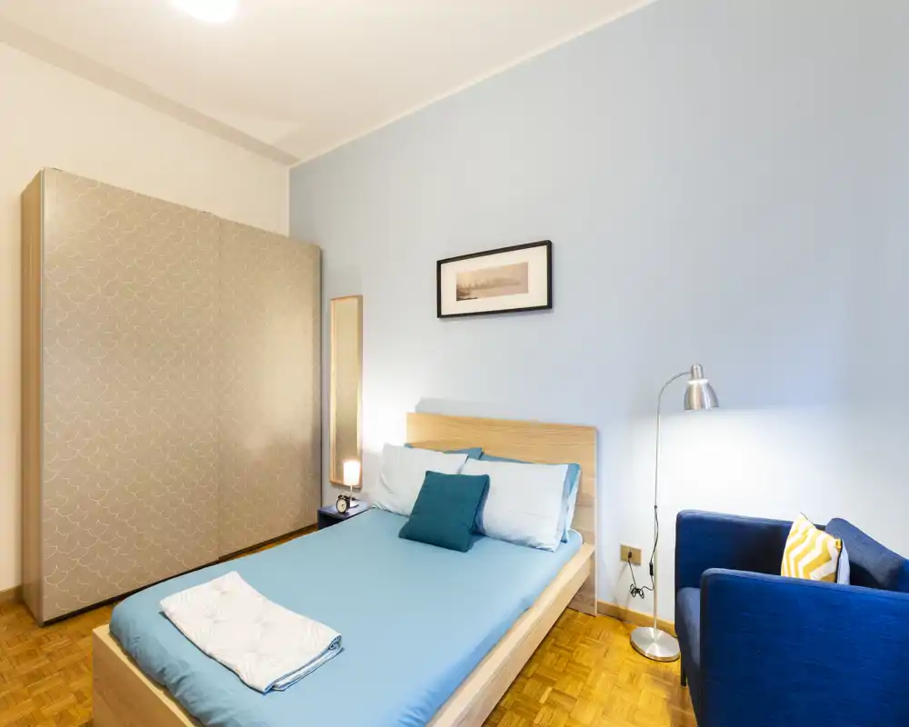 Bright, modern bedroom with a double bed dressed in blue linens, a patterned wardrobe, and a cozy navy armchair — ideal for a comfortable stay.