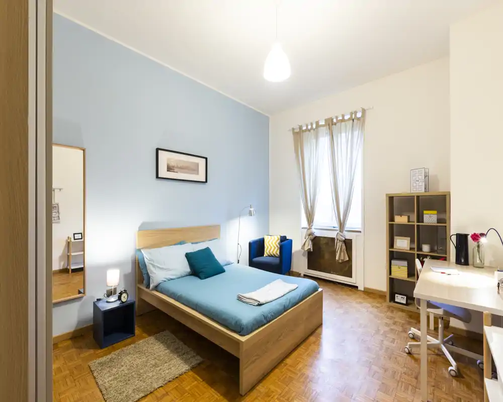 Bright, well-staged bedroom with a wooden double bed, blue accent wall, armchair and a work desk — ideal for a comfortable stay.