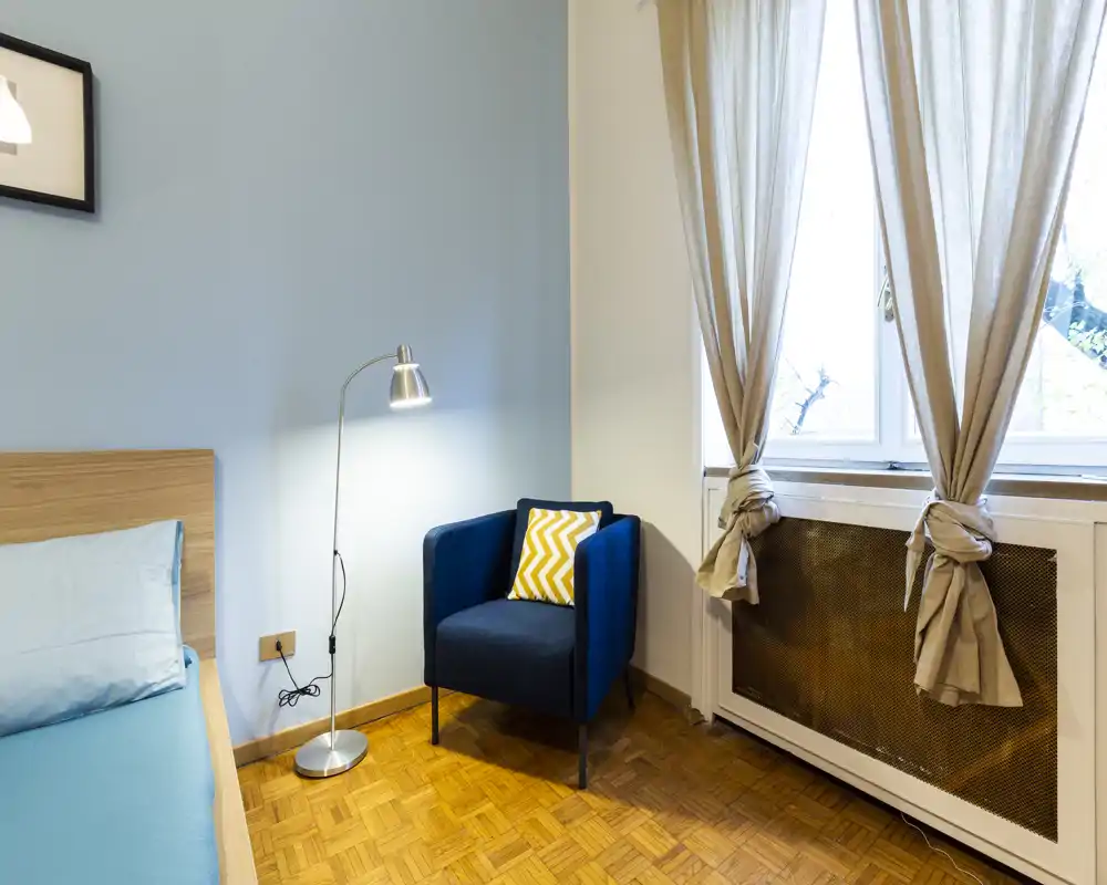 Bright, neatly styled bedroom corner with a visible bed, a blue upholstered armchair with a patterned cushion, a floor lamp, and tied-back curtains letting in natural light.