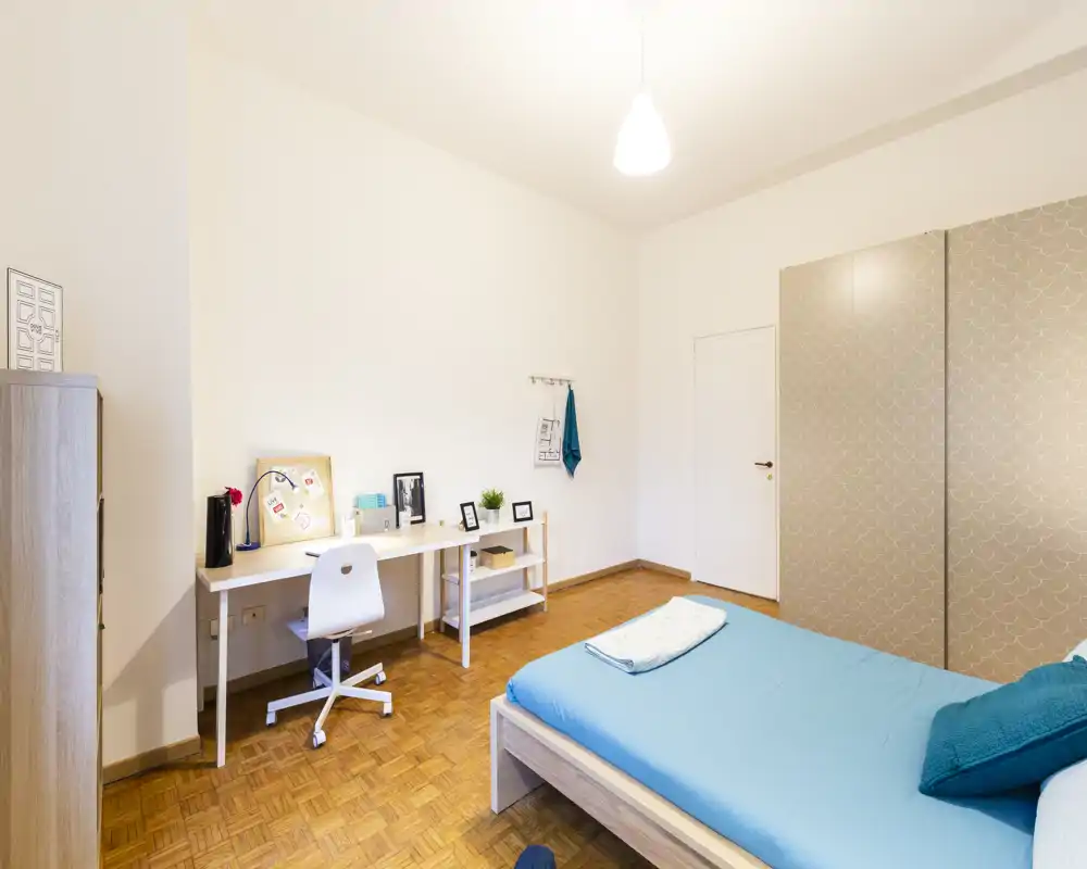 Bright, tidy bedroom with a double bed dressed in blue linens, a workspace with desk and chair, and a large wardrobe — ideal for a comfortable stay.
