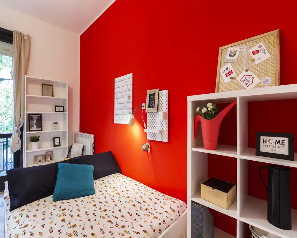 Bright, stylish bedroom with a bold red accent wall, well-made bed with patterned duvet and decorative cushions, shelving and natural light from a nearby window.