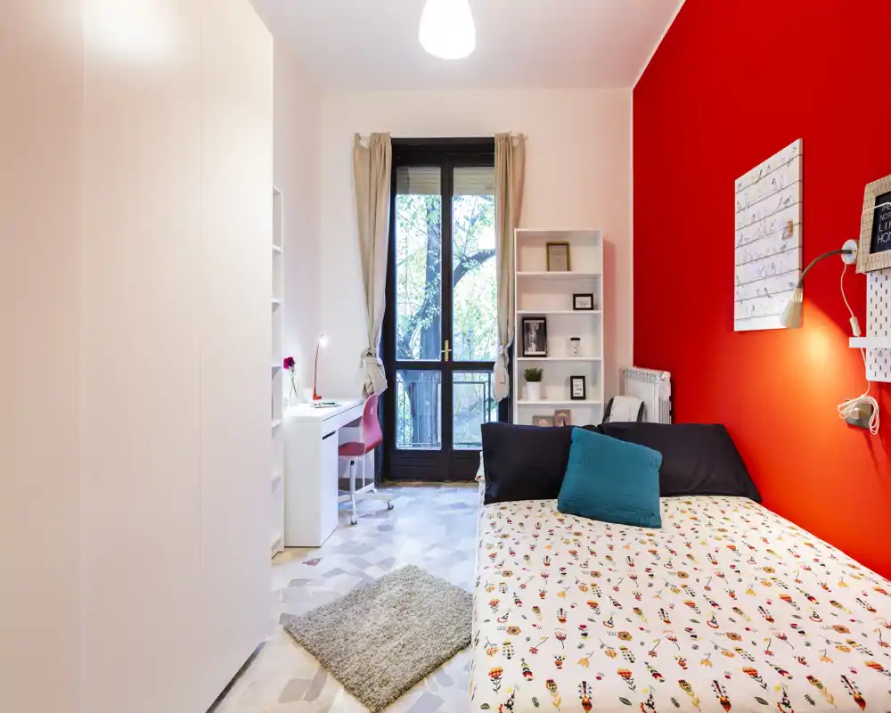Bright, modern bedroom with a double bed against a red accent wall, decorative bedding and a teal cushion, desk by the window and built-in shelving — ideal for a student or single occupant.
