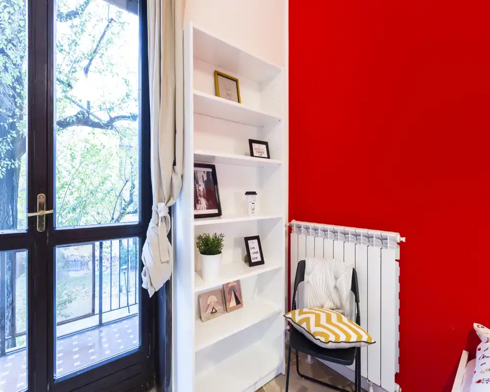 Small bedroom corner with a large window and balcony access, a tall white shelving unit with framed photos and plants, and a chair with a chevron cushion against a vivid red wall.