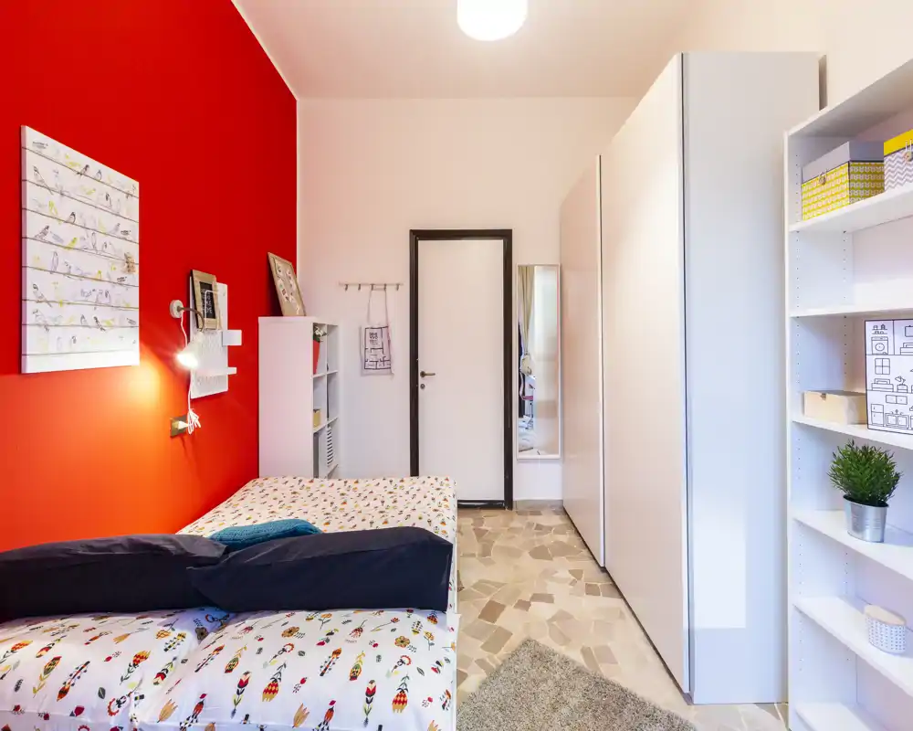 Bright, modern single bedroom with colorful bedding, a bold red accent wall, wardrobe and shelving — tidy and well lit.