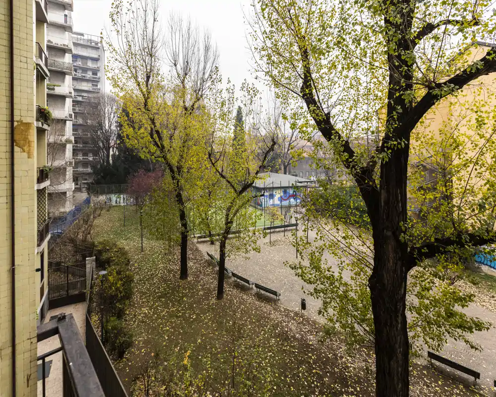 View of a communal outdoor courtyard with trees, benches and a small fenced play or sports area; autumn leaves cover the ground and surrounding apartment buildings are visible.