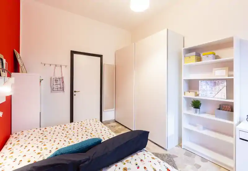 Camera da letto luminosa e ordinata con letto matrimoniale, armadio e mensole; la biancheria colorata e la parete rossa creano un ambiente caldo e moderno.