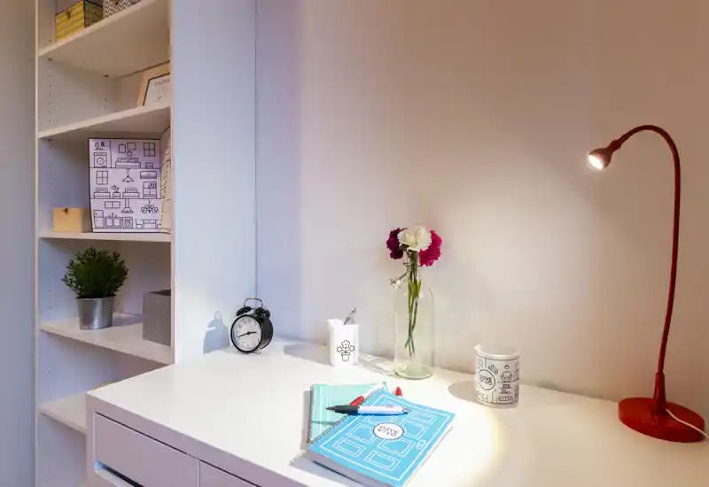 Angolo scrivania luminoso e ordinato con tocchi decorativi: lampada da tavolo rossa, vaso con fiori, sveglia e quaderni su una superficie bianca vicino a scaffalatura aperta.