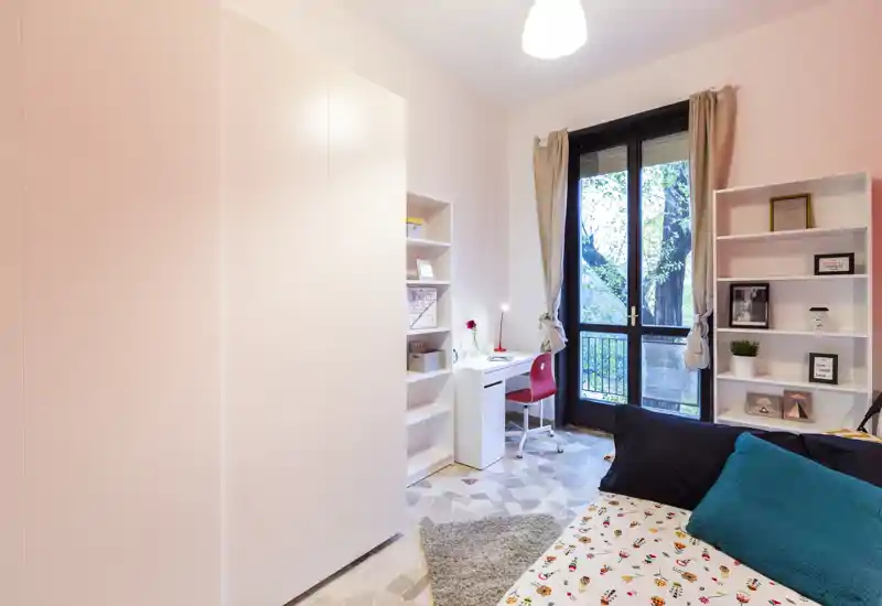 Camera da letto luminosa e moderna con copriletto a fantasia, cuscini accoglienti, scaffalature e una scrivania vicino alla finestra che offre luce naturale e una vista gradevole.