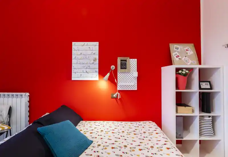 Camera da letto luminosa e di stile con copriletto fantasia, cuscini turchesi e una parete rossa vistosa. La lampada da comodino e lo scaffale offrono spazio e dettagli decorativi.