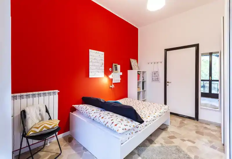 Camera singola luminosa e moderna con letto bianco, biancheria pattern e parete rossa d'accento. La stanza include un mobile a scaffali, una sedia con cuscini e una finestra che porta luce naturale — perfetta per un affitto accogliente.