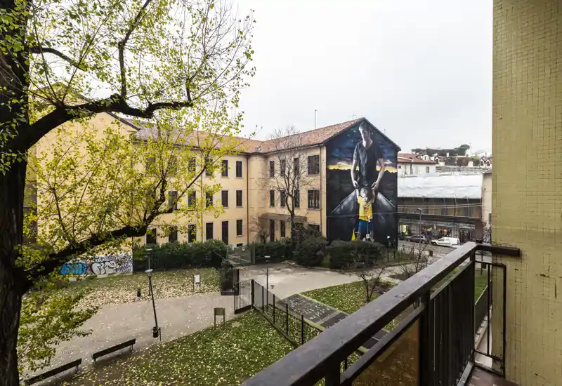 Vista da un balcone su un cortile con panchine, alberi e un grande murale sulla facciata dell'edificio di fronte.