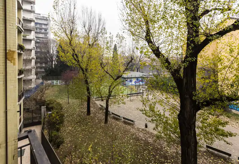Cortile condominiale con alberi maturi, panchine e un piccolo spazio pavimentato; foglie autunnali sul terreno e un edificio residenziale adiacente.