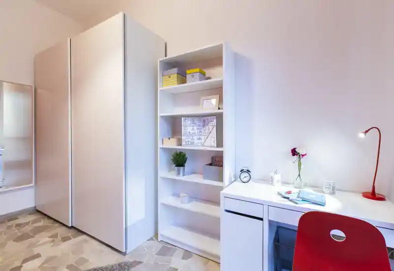 Angolo camera luminoso e moderno con armadio bianco, scaffale e scrivania ordinata con sedia rossa e lampada; ideale per una persona o studente.